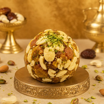 MM Mithaiwala Sugar Free Jardalu Laddu topped with pistachios on a decorative gold pedestal with festive brass decor behind.
